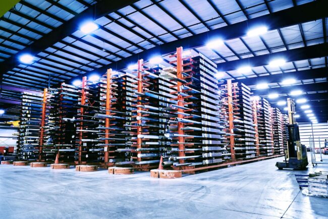 Marmon Keystone A large warehouse with tall, multi-level metal racks storing long tube pipe bars and metals. The well-lit space has a clean concrete floor and an organized supplychain layout. A forklift is visible on the right side, highlighting an efficient distributor environment.