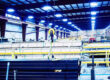 Marmon Keystone A brightly lit industrial warehouse interior with stacked tube pipes and metals, yellow safety railings, and a hoist system hanging from the ceiling, showcasing a busy supplychain distributor environment.